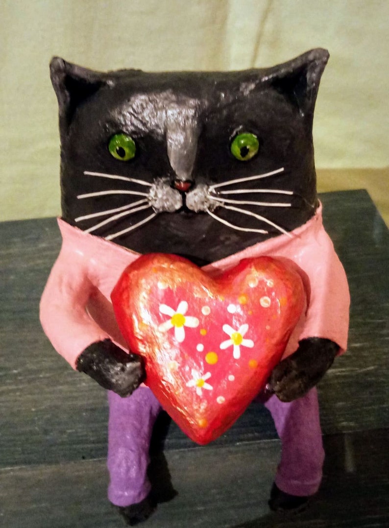 May include: A black cat figurine wearing a pink shirt and holding a red heart with white flowers. The heart is decorated with orange dots.