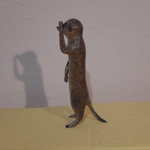 May include: A brown meerkat statue stands upright, shielding its eyes with one hand. The sculpture has a textured surface and a long tail. The meerkat is positioned on a yellow surface against a white wall.