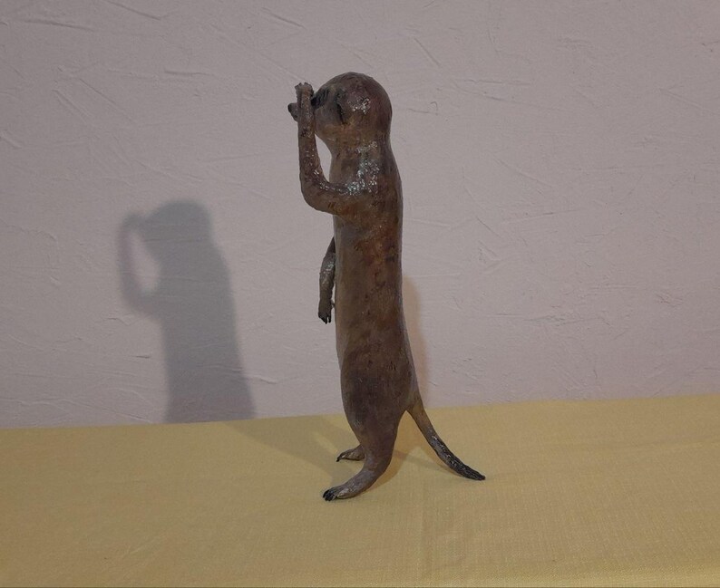 May include: A brown meerkat statue stands upright, shielding its eyes with one hand. The sculpture has a textured surface and a long tail. The meerkat is positioned on a yellow surface against a white wall.