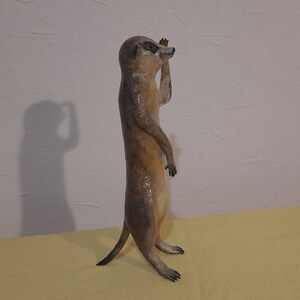 May include: A standing meerkat figurine with a light brown body and a darker brown head and tail. The meerkat is posed with one arm raised, as if looking out. The figurine is on a yellow surface, with a shadow cast on the wall behind it.