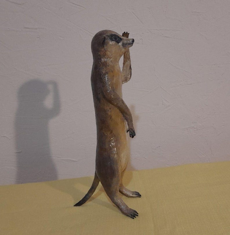 May include: A standing meerkat figurine with a light brown body and a darker brown head and tail. The meerkat is posed with one arm raised, as if looking out. The figurine is on a yellow surface, with a shadow cast on the wall behind it.