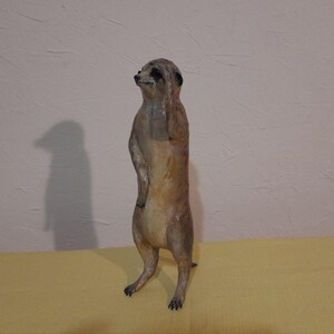 May include: A handcrafted meerkat figurine in shades of brown and tan, standing upright with one paw raised to its head. The meerkat has black markings around its eyes and a detailed, textured surface. The figurine is on a yellow surface.