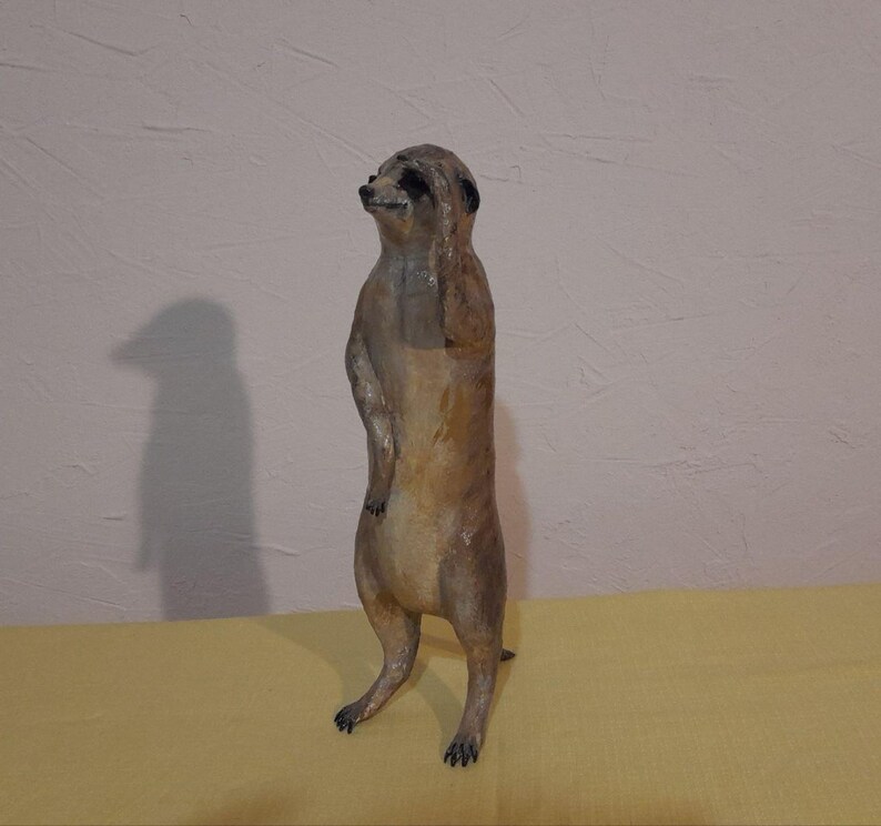 May include: A handcrafted meerkat figurine in shades of brown and tan, standing upright with one paw raised to its head. The meerkat has black markings around its eyes and a detailed, textured surface. The figurine is on a yellow surface.