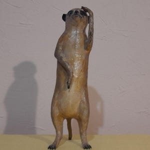May include: A standing meerkat figurine with a light brown body and black ears. The meerkat has one arm raised to its head, as if in thought. The figurine is on a yellow surface.