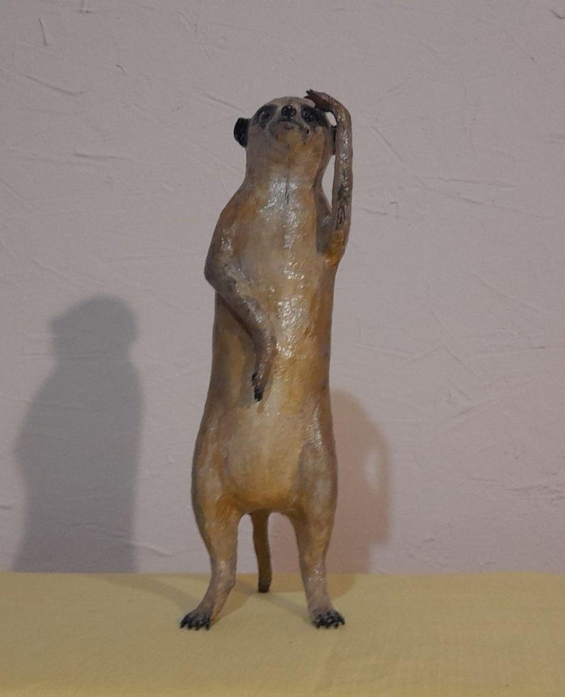 May include: A standing meerkat figurine with a light brown body and black ears. The meerkat has one arm raised to its head, as if in thought. The figurine is on a yellow surface.