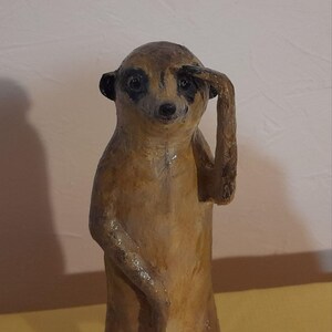 May include: A handcrafted meerkat figurine in a standing pose, shielding its eyes with one arm. The sculpture is primarily brown with black accents around the eyes and ears. The meerkat appears to be made of a textured material.