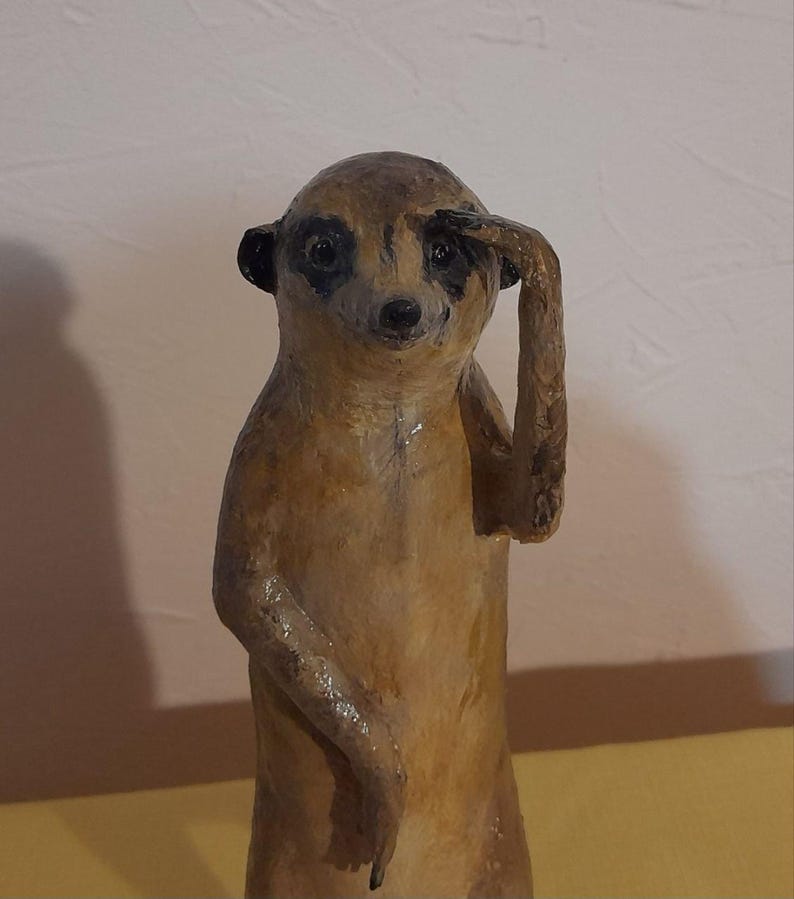 May include: A handcrafted meerkat figurine in a standing pose, shielding its eyes with one arm. The sculpture is primarily brown with black accents around the eyes and ears. The meerkat appears to be made of a textured material.