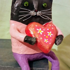 May include: A black cat figurine wearing a pink shirt and purple pants holds a red heart with white flowers and orange dots.
