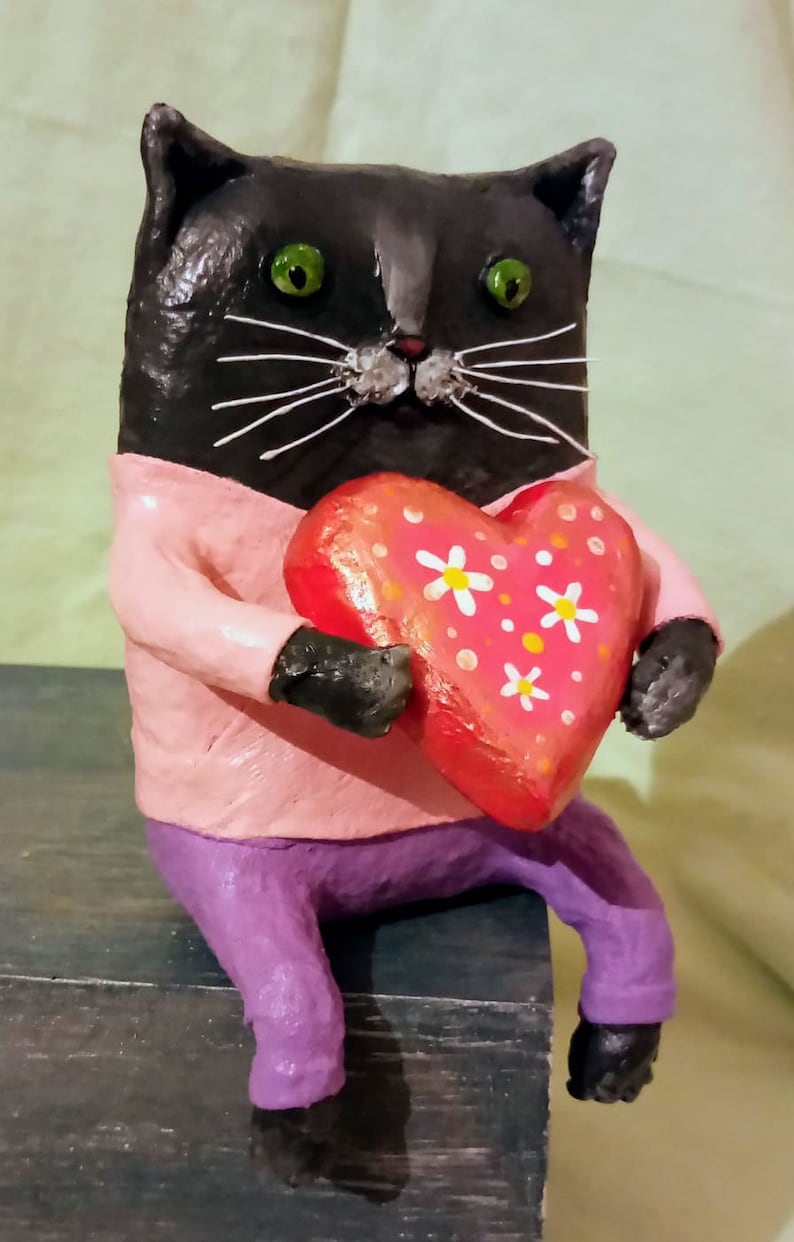 May include: A black cat figurine wearing a pink shirt and purple pants holds a red heart with white flowers and orange dots.
