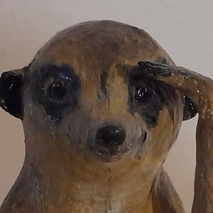 May include: A handcrafted meerkat figurine with a brown and black color scheme. The meerkat is depicted with its arm raised to its head, as if saluting. The figurine has a glossy finish and detailed facial features.