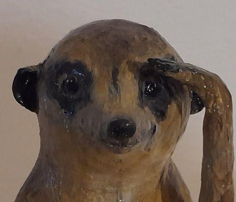 May include: A handcrafted meerkat figurine with a brown and black color scheme. The meerkat is depicted with its arm raised to its head, as if saluting. The figurine has a glossy finish and detailed facial features.