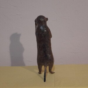 May include: A brown meerkat figurine standing upright, with its arms held close to its body. The sculpture has a textured surface and a small tail. The meerkat is positioned on a yellow surface against a light pink wall.