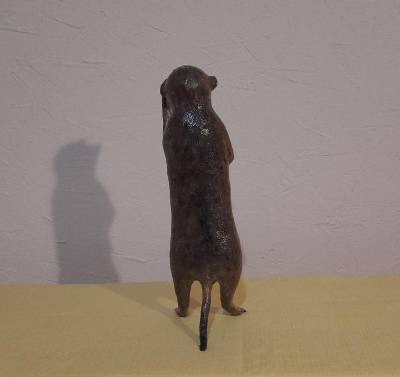 May include: A brown meerkat figurine standing upright, with its arms held close to its body. The sculpture has a textured surface and a small tail. The meerkat is positioned on a yellow surface against a light pink wall.