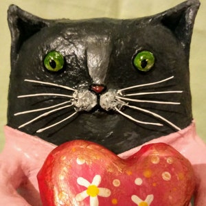 May include: A black cat figurine with green eyes and white whiskers holds a pink heart-shaped object with white daisies and orange dots.