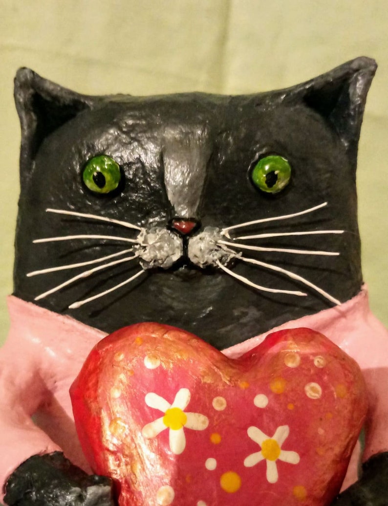 May include: A black cat figurine with green eyes and white whiskers holds a pink heart-shaped object with white daisies and orange dots.