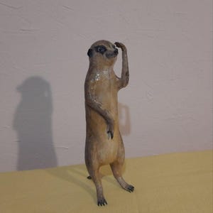 May include: A standing meerkat sculpture with a light brown body and dark markings around the eyes and ears. The meerkat is holding its right arm up to its head, as if shielding its eyes. The sculpture is on a yellow surface.
