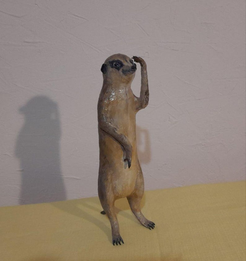May include: A standing meerkat sculpture with a light brown body and dark markings around the eyes and ears. The meerkat is holding its right arm up to its head, as if shielding its eyes. The sculpture is on a yellow surface.