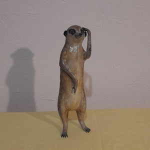 May include: A handcrafted meerkat figurine, standing upright with one arm raised to its forehead. The sculpture is painted in shades of brown and tan, with black accents on the eyes and ears. The meerkat is posed as if it is shielding its eyes from the sun.