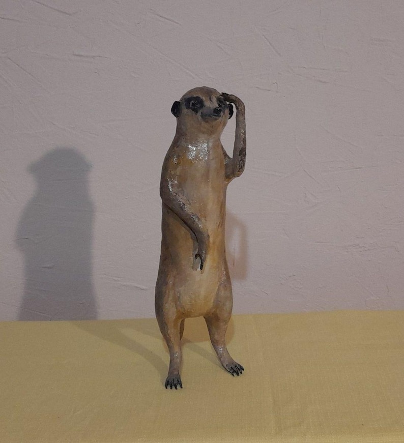 May include: A handcrafted meerkat figurine, standing upright with one arm raised to its forehead. The sculpture is painted in shades of brown and tan, with black accents on the eyes and ears. The meerkat is posed as if it is shielding its eyes from the sun.
