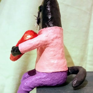 May include: A black cat figurine wearing a pink shirt and purple pants, holding a red heart in its paws. The cat is sitting on a wooden surface.