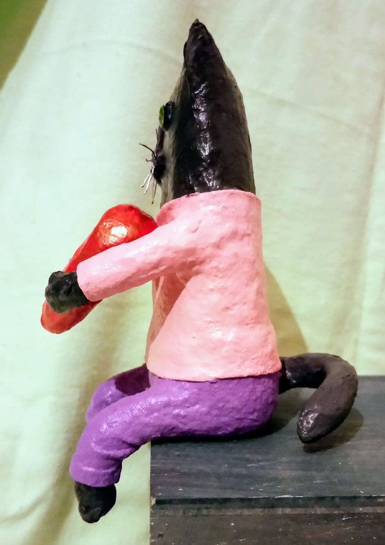 May include: A black cat figurine wearing a pink shirt and purple pants, holding a red heart in its paws. The cat is sitting on a wooden surface.