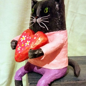 May include: A black cat figurine wearing a pink shirt and purple pants holds a red heart with white flowers. The cat is sitting on a wooden surface.