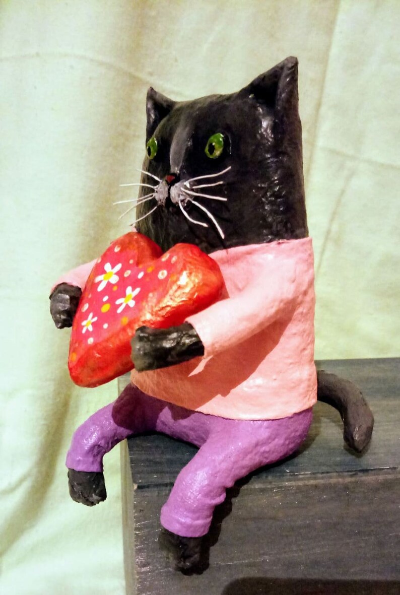 May include: A black cat figurine wearing a pink shirt and purple pants holds a red heart with white flowers. The cat is sitting on a wooden surface.
