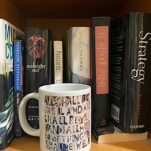 May include: White ceramic mug with a colorful collage of text that reads "All shall be well and all shall be well and all manner of thing shall be well". The mug is sitting on a shelf with a variety of books behind it.