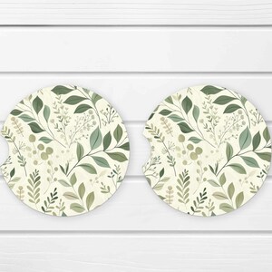 May include: Two round car coasters with a botanical design. The coasters feature a cream background with a pattern of green leaves and sprigs. One coaster has a notch for easy removal.