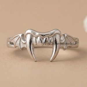 May include: A silver ring with a vampire teeth design. The ring features detailed fangs and bat wing accents. The ring is displayed on a neutral background with white flowers.