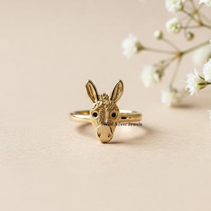 May include: A gold donkey head ring with black eyes. The ring is on a light brown surface.