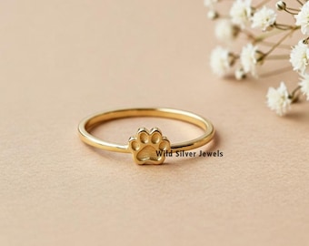 Puede incluir: Un anillo dorado con un diseño de huella de pata. El anillo está sobre un fondo marrón claro.