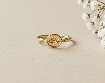 May include: A gold-colored ring with a circular face featuring a detailed fish design. The ring is set against a neutral background with white flowers, highlighting the jewelry's elegant simplicity. The ring is a delicate piece of jewelry.