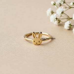 May include: A gold-colored ring with a bunny rabbit design. The ring is on a light brown surface.
