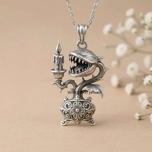 Könnte beinhalten: Ein silberner Anhänger mit einer fleischfressenden Pflanze mit scharfen Zähnen, die eine brennende Kerze in einem dekorativen Halter hält. Der Sockel ist mit Blumenmustern verziert. Der Text Wild Silver jewels ist am Stängel zu sehen.