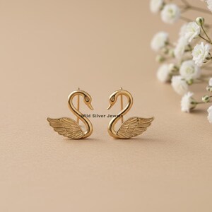 May include: A pair of gold-colored swan earrings. Each earring features a swan design with detailed wings and a curved neck. The earrings are set against a neutral background with white flowers.