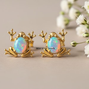 Puede incluir: Pendientes de botón dorados con forma de rana. Cada rana presenta una piedra de ópalo ovalada con colores iridiscentes. Los pendientes están sobre un fondo suave con flores blancas. El texto "Wild Silver Jewels" es visible.