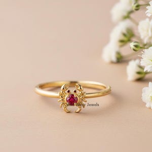 Pode incluir: Anel dourado com design de caranguejo, com uma pedra preciosa vermelha central e duas pedras vermelhas menores. O anel está sobre um fundo neutro, com flores brancas ao fundo.