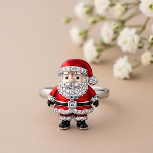 May include: A silver ring featuring a Santa Claus figure. The Santa is depicted with a red coat and hat, a white beard, and is embellished with sparkling crystals. The ring is a festive holiday accessory.