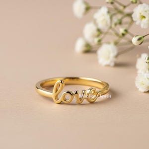 May include: A gold ring featuring the word "love" in a cursive font. The ring is set against a neutral background with white flowers. The ring is a delicate piece of jewellery.
