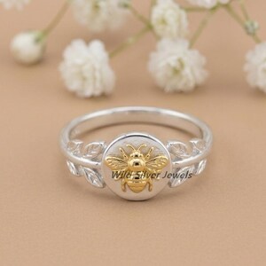 May include: A silver ring with a gold bee design. The ring features a circular gold bee emblem in the center, surrounded by a silver band with leaf accents. The background is a light brown color with white flowers.