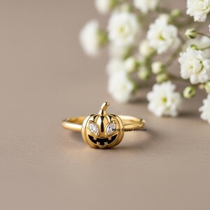 May include: A gold-colored ring featuring a carved pumpkin design with two clear gemstones for eyes and a carved mouth. The ring is set against a neutral background with white flowers.