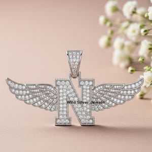 May include: A silver pendant in the shape of the letter "N" with encrusted clear gemstones, flanked by detailed wings. The pendant is set against a soft, neutral background, with the text "Wild Silver Jewels" visible.