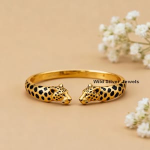 May include: A gold-coloured bangle bracelet with two leopard heads facing each other. The leopards have black spots and detailed facial features. The bracelet is set against a neutral background.