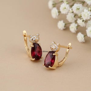 Puede incluir: Un par de pendientes dorados con forma de gato. La cabeza del gato está incrustada con pequeñas piedras transparentes. El cuerpo del pendiente presenta una gran piedra roja oscura en forma de lágrima. Fondo neutro.