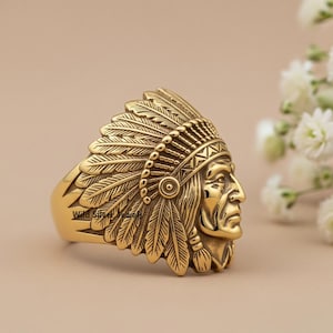 May include: A gold-colored ring featuring a detailed Native American chief headdress design. The ring showcases a profile of a Native American chief with intricate feather detailing. The ring is set against a neutral background.