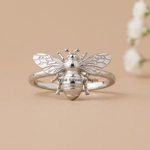 May include: A silver-colored bee ring. The ring features a detailed bee design with textured wings and body. The ring is set against a neutral background, with a sprig of white flowers in the background.
