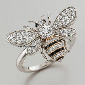 May include: A silver ring with a bee design. The bee is covered in clear sparkling stones and has black and gold stripes. The words "Wild Silver Jewels" are visible on the ring.