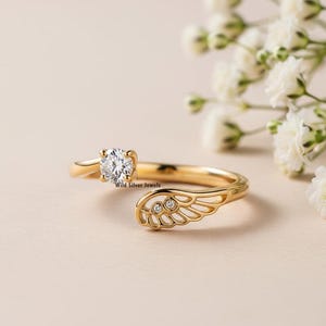 May include: A gold-colored ring with a clear, round gemstone and a wing-shaped design. The ring features three smaller gemstones on the wing. The text "Wild Silver Jewels" is visible on the ring.
