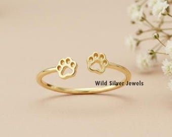Puede incluir: Un anillo de color dorado con dos diseños de huellas de pata. El anillo es delicado y simple, perfecto para el uso diario. "Wild Silver Jewels" está impreso en el fondo.
